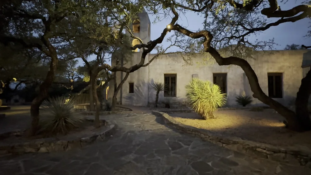 The courtyard of the Old World Huerta at dusk: the stone-edged bed under the live oaks, the lit mission wall beyond.