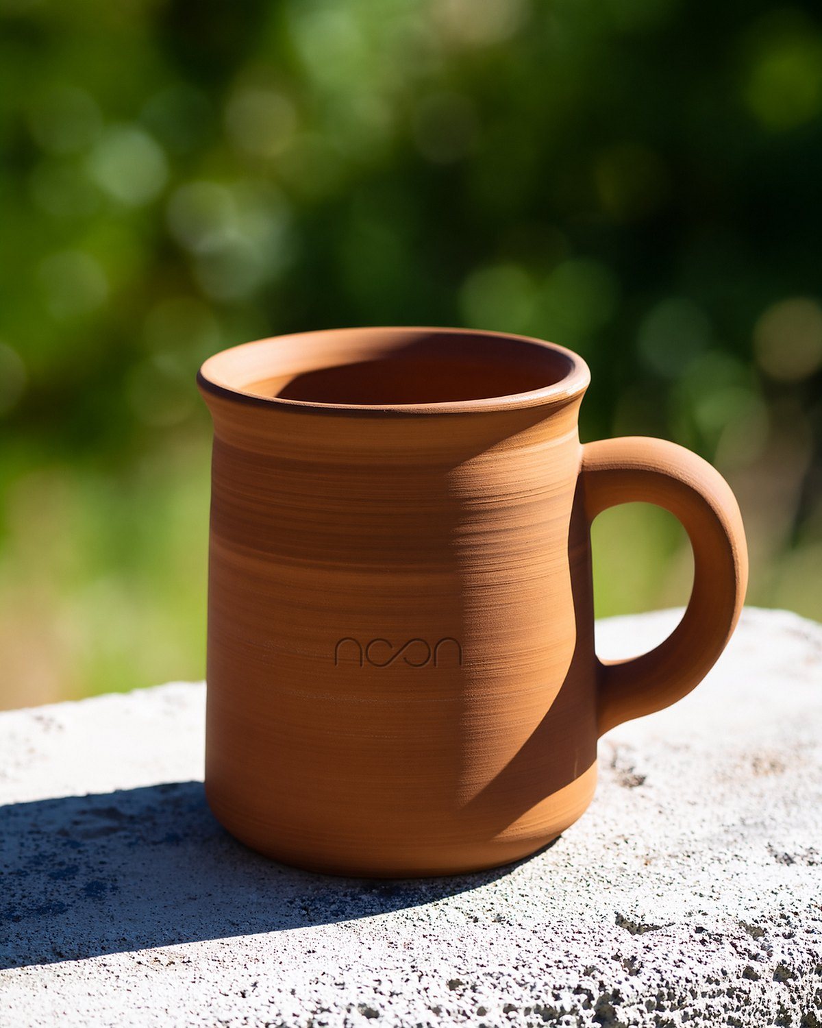 A handmade terracotta mug on a concrete ledge in the sun, the word noon pressed small into the clay on the side facing out