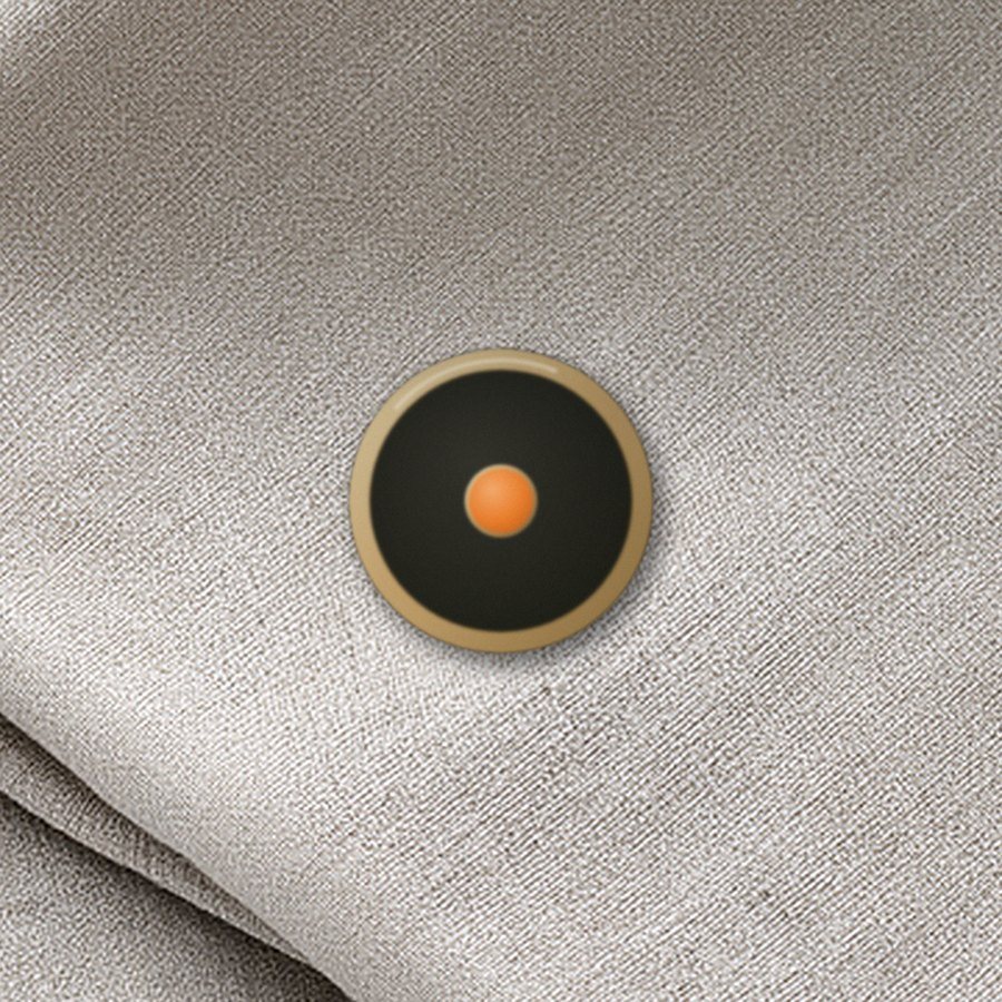 A small round enamel pin on linen: a brass ring, a dark field, an ember dot