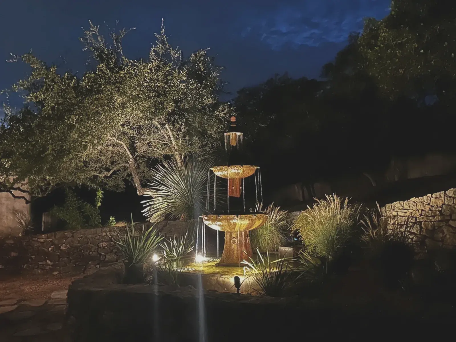 A tiered steel fountain running at nightfall, uplit among native grasses, yucca, and live oaks