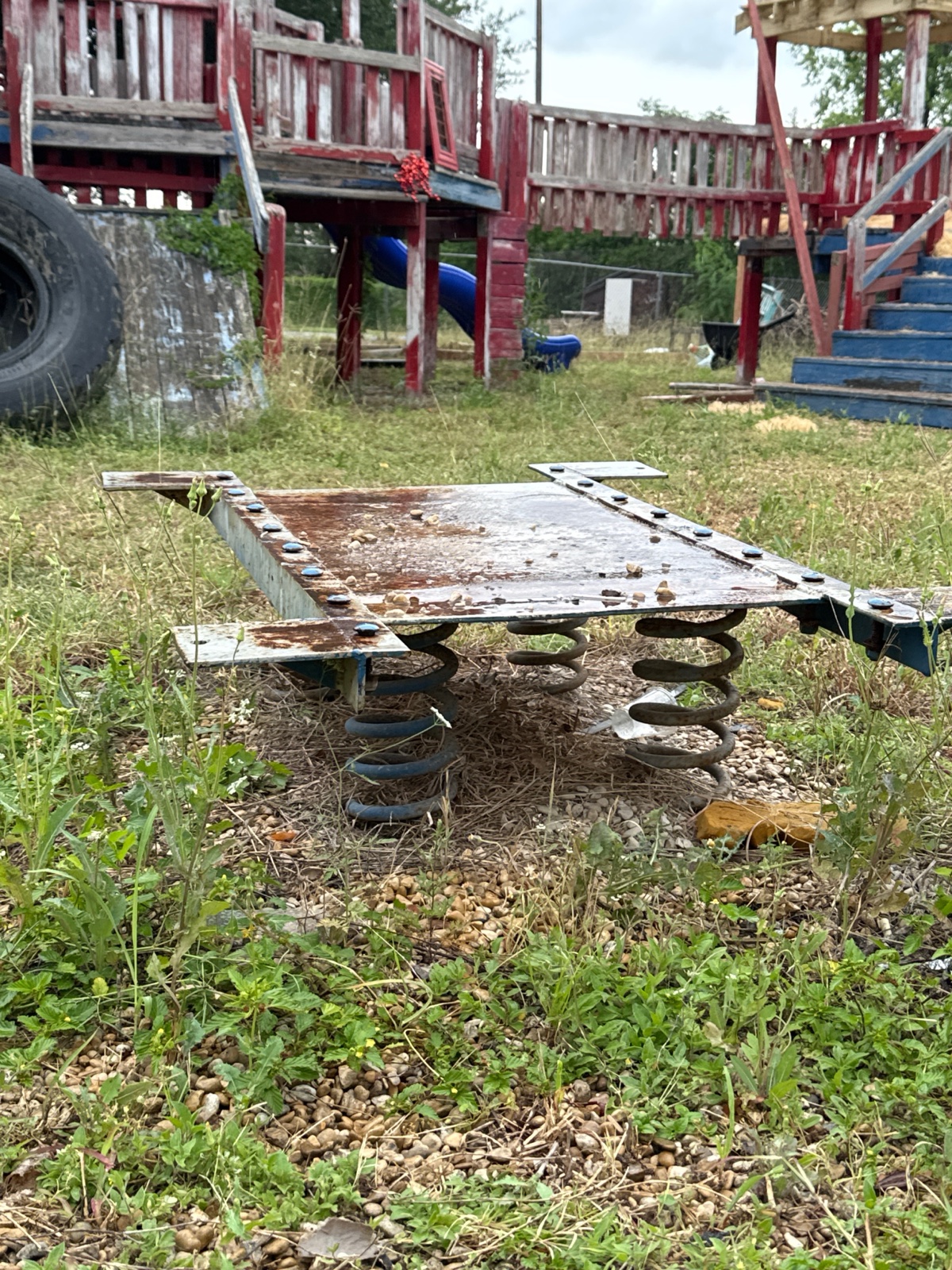 A rusted, broken playground spring-toy in overgrown weeds behind the building.