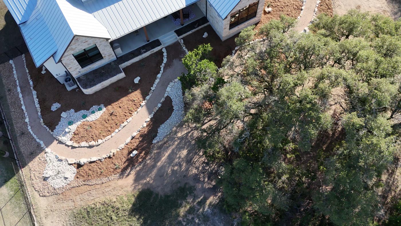 Aerial view of the Dry-Creek Xeriscape — decomposed-granite paths and a river-rock dry creek around a metal-roof Hill Country home.