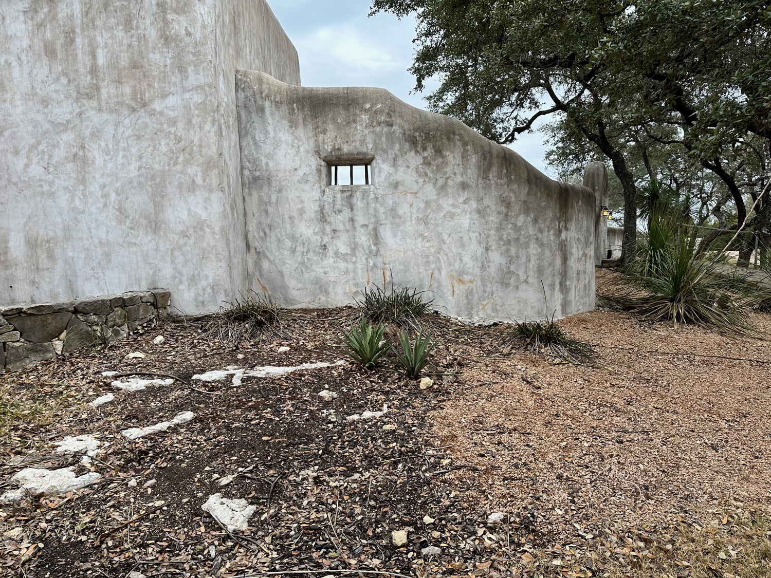 Bare, dry, rocky Hill Country ground — caliche and limestone — at the foot of a mission-styled wall, before planting.