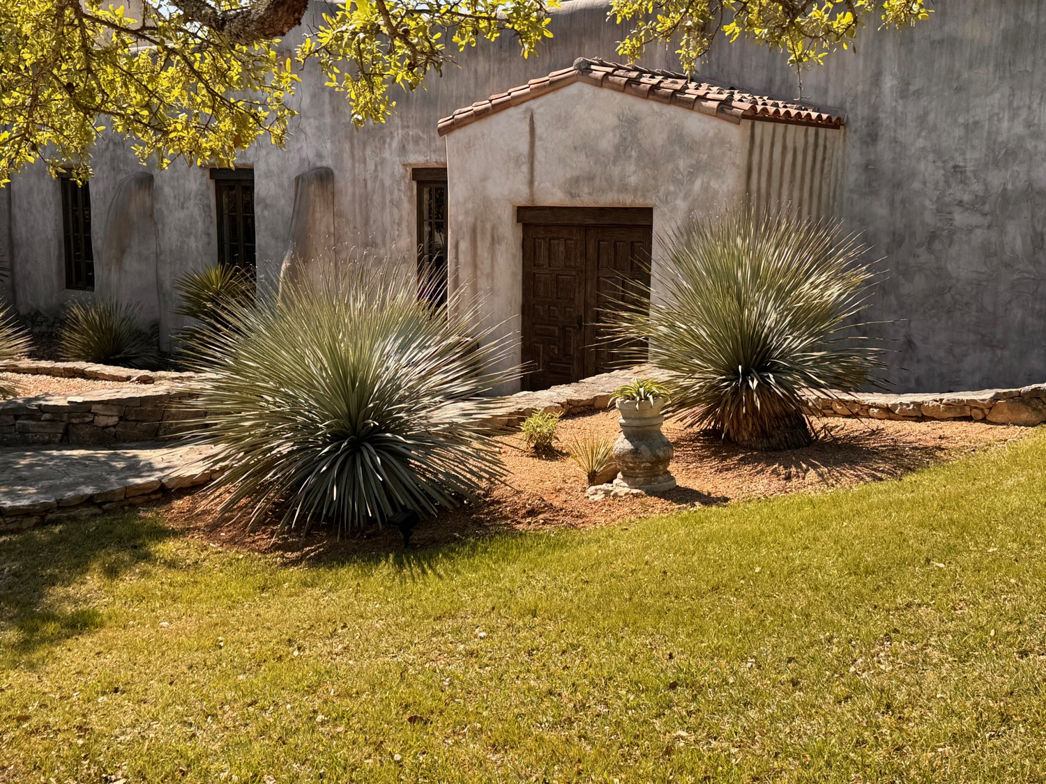 A tile-roofed mission-styled building with stone-edged beds of sotol and a stone urn planter.