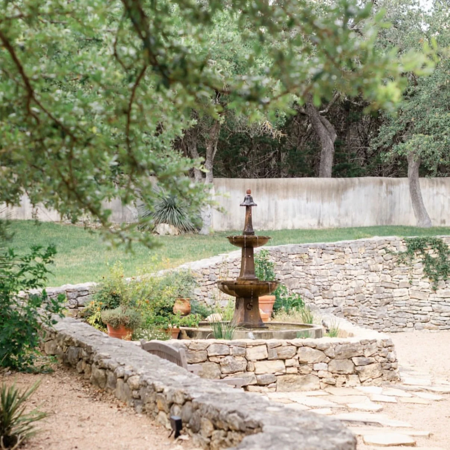 The Huerta garden — a tiered fountain in a stone-walled forecourt, terraced beds and potted herbs under green live oaks.
