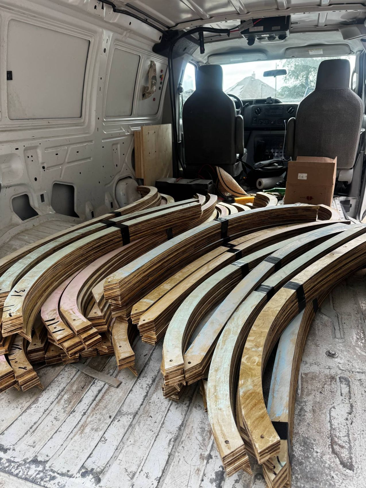Dozens of curved laminated birch formwork ribs, numbered in marker and strapped in bundles, filling the cargo floor of a work van.